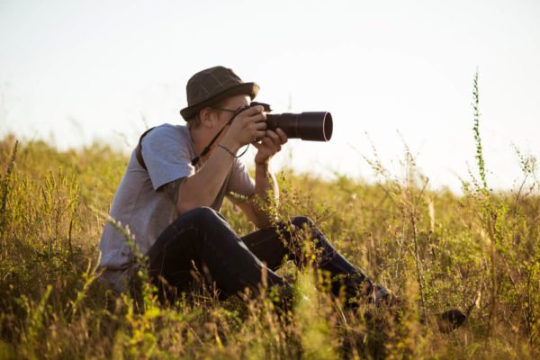 young-male-photographer-hat-taking-picture-sitting-field (1)